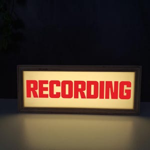 Könnte beinhalten: Ein rechteckiges Schild mit Holzrahmen und dem Wort "RECORDING" in fetten roten Buchstaben. Das Schild hat einen warmen, beleuchteten Hintergrund, was darauf hindeutet, dass es von innen beleuchtet wird. Das Design ist einfach und funktional.