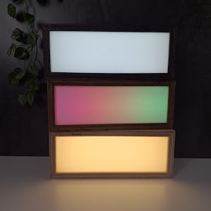 May include: Three rectangular LED light panels stacked on top of each other. The top panel is white, the middle panel has a pink to green gradient, and the bottom panel is yellow. Each panel is framed in a dark wood.