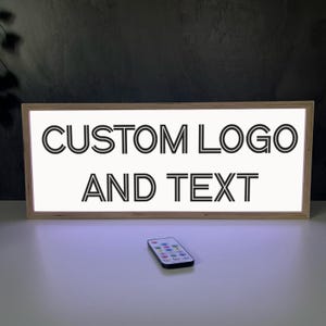 May include: A rectangular light box with a wooden frame displays the text "CUSTOM LOGO AND TEXT" in black. A small remote control with multiple buttons sits below the light box. The light box is on a white surface.