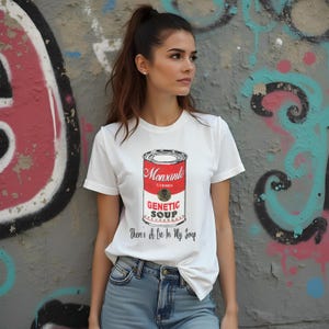 May include: White t-shirt featuring a graphic of a red and white can of soup. The can has the text "Monsanto" and "Genetic Soup" on it. The text "There's A Lie In My Soup" is printed below the can.