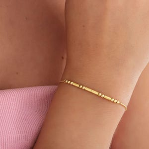 Könnte beinhalten: Ein filigranes goldenes Armband mit kleinen Perlen und zylindrischen Stäben. Das Armband wird am Handgelenk getragen und unterstreicht sein minimalistisches Design und seinen warmen, metallischen Farbton. Das Armband ist etwa 18 cm lang.