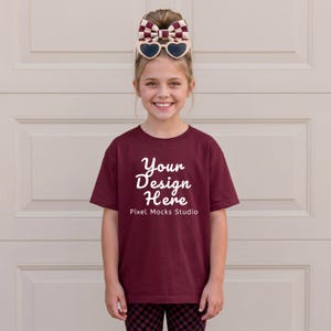 Può includere: Una persona giovane indossa una maglietta bordeaux con la scritta "Your Design Here" in bianco. La maglietta è abbinata a pantaloni a scacchi neri e rossi. Un fiocco con occhiali da sole a forma di cuore è tra i capelli.