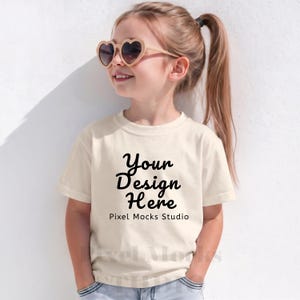 Może przedstawiać: Młoda osoba w kremowym t-shircie z czarnym napisem "Your Design Here". Osoba ta nosi również okulary przeciwsłoneczne w kształcie serca z jasnobrązową oprawką. Tło to biała ściana.