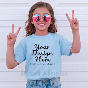 Op de afbeelding: Een jong meisje draagt een lichtblauw t-shirt met de tekst "Your Design Here" in zwart schrift. Ze draagt ook een ronde, roze getinte zonnebril en maakt een vredesteken met haar vingers.