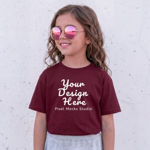Può includere: Una maglietta bordeaux con la scritta "Your Design Here" in stile scritto a mano, con "Pixel Mocks Studio" sotto. La modella indossa occhiali da sole rotondi con lenti rosa.