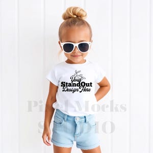 May include: A white t-shirt with short sleeves and the text "Your StandOut Design Here" printed on the front. A young girl is wearing the shirt and blue denim shorts. She is wearing white sunglasses.