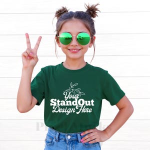 May include: A young person wearing a dark green t-shirt with the text "Your StandOut Design Here" in white script. The person is also wearing round green sunglasses and blue jeans. The person is making a peace sign with their fingers.
