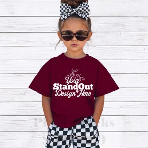 Può includere: Una persona giovane indossa una maglietta bordeaux con la scritta bianca "Your StandOut Design Here". La persona indossa anche pantaloncini a scacchi bianchi e neri, occhiali da sole e un fiocco abbinato. Lo sfondo è un muro di legno bianco.