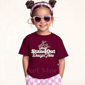 Può includere: Un bambino indossa una maglietta bordeaux con la scritta bianca "Your StandOut Design Here". Il bambino indossa anche occhiali da sole rosa a forma di cuore e una fascia a scacchi rosa e bianca.