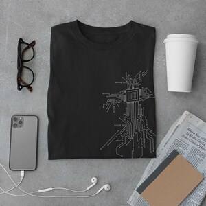 Könnte beinhalten: Ein schwarzes T-Shirt mit einem weißen Schaltkreisdesign. Das Shirt ist gefaltet und liegt flach auf einer grauen Oberfläche. Auf dem Bild sind auch eine Brille, ein Smartphone, eine Tasse Kaffee, ein Notizbuch und eine Zeitung zu sehen.