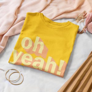 Könnte beinhalten: Ein gelbes T-Shirt mit dem Aufdruck "oh yeah!" in rosa und gelben Buchstaben.