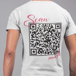 Könnte beinhalten: Weißes T-Shirt mit einem schwarz-weißen QR-Code und dem Text "Scan mich" in rosa Kursivschrift.