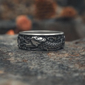 May include: A silver ring with an intricate design of a fish, set against a blurred background of trees and foliage.