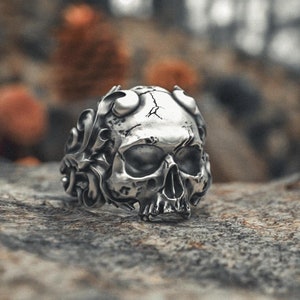 May include: A silver skull ring with intricate details, featuring a skull with a floral design around the head. The ring is sitting on a textured rock surface.