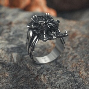 May include: A silver ring with a detailed, snarling monster head design. The ring is on a textured, gray surface.
