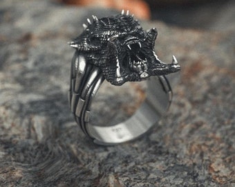 May include: A silver ring featuring a detailed, snarling monster head design. The ring is resting on a textured, grey surface.