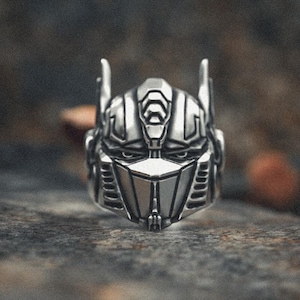 Puede incluir: Un anillo de plata con un diseño detallado de una cabeza de robot. La cabeza del robot tiene una visera negra y una cara plateada.