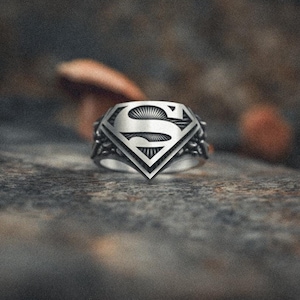 Può includere: Un anello d'argento con un design a logo Superman. L'anello è su una superficie testurizzata di colore marrone.