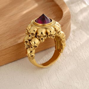 May include: Gold-coloured ring with a skull design around the band. The ring is topped with a large, faceted, deep red gemstone. The ring is displayed on a wooden surface.