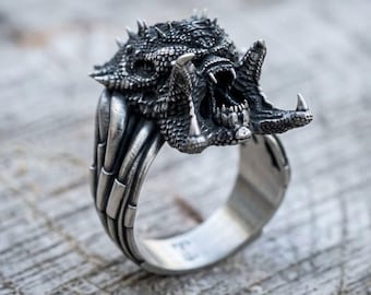 May include: A silver-toned ring featuring a detailed, textured creature head with an open mouth, sharp teeth, and spikes. The band of the ring is designed with vertical, rib-like segments that extend from the base of the creature head downward.