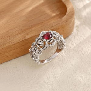May include: A silver ring with a heart-shaped, red gemstone at the center. The ring features an intricate, openwork design with floral and geometric patterns. The ring is set against a light-colored background.
