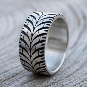 May include: A wide silver-toned metal ring featuring a detailed, recessed tire tread pattern that wraps around the entire exterior band. The design includes deep black grooves contrasting with the brushed metallic surface of the tire tread design.
