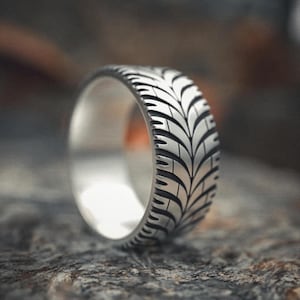 May include: A silver ring with a tire tread pattern. The ring is made of a shiny metal and has a detailed design that resembles the tread of a tire. The ring is shown on a textured, gray surface.