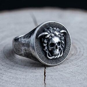 May include: A silver ring with a skull design. The ring features a detailed skull within a circular frame with a Greek key pattern. The band has a textured, hammered appearance, giving it a rustic look.
