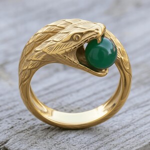 May include: A gold-toned ring featuring a detailed snake design with textured scales. The snake's head is curved, with its mouth open to hold a smooth, round, dark green bead. The band is solid and polished, forming a continuous loop around the finger.