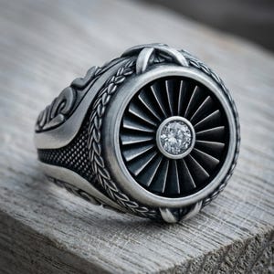 Könnte beinhalten: Ein silberfarbener Ring mit einer kreisförmigen Vorderseite, die an eine Strahltriebwerksturbine mit strahlenförmigen Schaufeln erinnert. Ein einzelner, klarer, rund geschliffener Edelstein ist in der Mitte gefasst. Die Ringschiene zeigt komplexe Gravuren.