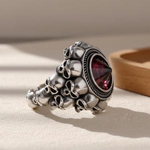 May include: Silver ring with a design of small skulls and a large, faceted, red gemstone. The ring has a detailed, gothic design with a textured band and a prominent central stone, ideal for a bold statement.
