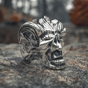Può includere: Un anello a teschio argentato dettagliato con intagli intricati, su uno sfondo naturale sfocato. L'anello presenta una superficie strutturata e una struttura facciale dettagliata, che mostra un'estetica gotica o motociclistica.