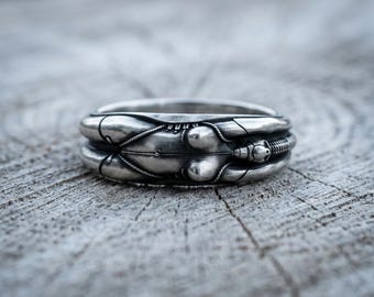 May include: A silver ring featuring a detailed, sculptural design of a beetle with a segmented body, rounded wing covers, and a small head with antennae. The metal has an oxidized finish that highlights the intricate lines and textures.