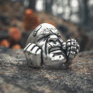 May include: A silver ring featuring a detailed, angry face with clenched fists. The ring has a textured surface and the word "HOOLIGAN" is visible on the mouth. The ring is set against a blurred natural background.