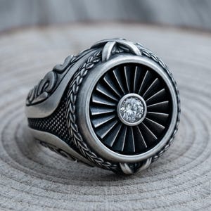 May include: A silver ring featuring a detailed jet engine design. The ring has a central fan blade motif with a sparkling, round gemstone at its center. Intricate patterns and textures adorn the band, creating a bold and unique accessory.