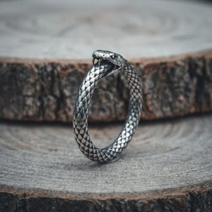 May include: A silver-toned metal ring shaped like a coiled snake biting its own tail. The band features a detailed, textured scale pattern across its entire surface, with the snake's head clearly defined at the top, showing visible fangs and eyes.