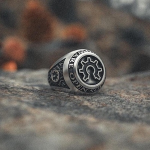 Op de afbeelding: Een zilveren ring met een zwart tandwielontwerp en de woorden "We Are Open Source" gegraveerd rond het tandwiel.