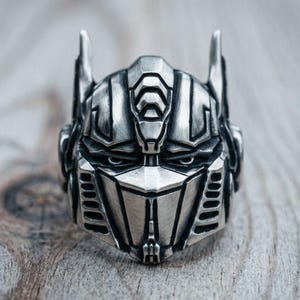 Könnte beinhalten: Ein silberfarbener Metallring in Form eines Roboterhelms mit spitzem Kamm, vertikalen Lüftungsschlitzen an den Seiten und einer geometrischen Frontplatte. Die Oberfläche hat ein gebürstetes Finish mit dunklen Vertiefungen für Details.
