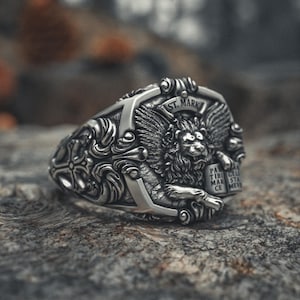 May include: A detailed silver ring featuring a winged lion design, with the text "ST. MARK" above the lion. The ring has intricate scrollwork and a cross design on the band. The ring is displayed on a textured, gray surface.