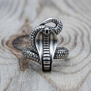 Può includere: Un anello color argento con un design dettagliato a forma di cobra. Il serpente è avvolto con il cappuccio spiegato e la bocca aperta, mostrando squame testurizzate sul corpo e un motivo liscio e segmentato al centro del cappuccio del cobra.