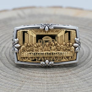 May include: A rectangular ring featuring a detailed gold-toned relief of The Last Supper scene. The central golden image is framed by a polished silver-toned border with ornate floral-inspired accents at the top, bottom, and sides of the band.