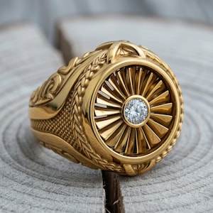 May include: A gold-toned ring featuring a circular face with a radial, turbine-like pattern and a single, clear, round-cut gemstone set in the center. The band is decorated with intricate, raised leaf-like engravings along the sides.