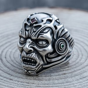 May include: A silver-colored ring with a detailed, sculpted design of a zombie-like face. The ring features a red gemstone on the forehead and a green gemstone on the side. The ring has a gothic or fantasy theme.
