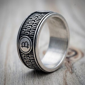 Peut inclure: Une bague en métal argenté présentant un symbole Bitcoin en relief à l'intérieur d'une bordure circulaire, entouré d'un motif répétitif de chiffres binaires 0 et 1 gravés sur l'anneau avec une finition sombre oxydée dans les creux.