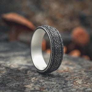 May include: A silver ring with an intricate floral pattern on the band. The ring has a white interior and is set against a blurred, textured background. The ring's design is detailed and ornate, suggesting a vintage or handcrafted style.