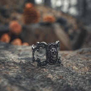 May include: A silver skull ring with intricate details and a black background. The ring is set against a blurred background of a forest.