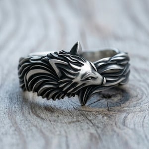 Puede incluir: Un anillo de metal plateado con un diseño detallado y esculpido de cabeza de lobo. La banda está elaborada con texturas de pelaje fluidas e intrincadas que envuelven el dedo, resaltando las orejas puntiagudas, el hocico y los ojos del lobo.