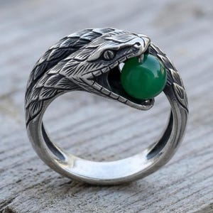 May include: A silver-toned metal ring featuring a detailed snake design that wraps around the finger. The snake's head is positioned at the top, with its mouth open, holding a smooth, round, vibrant green bead between its jaws.