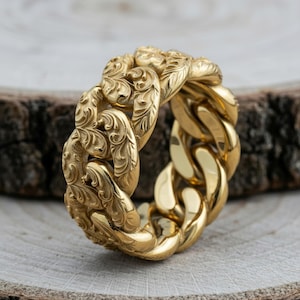 Puede incluir: Un anillo de color dorado con forma de cadena de eslabones gruesos. La superficie exterior de los eslabones metálicos presenta un intrincado patrón de volutas florales grabadas, mientras que el interior del anillo es liso y pulido con brillo.