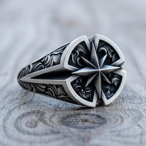May include: A silver-toned ring featuring a prominent eight-pointed star design at the center. The surface of the ring is decorated with intricate, swirling floral-like engravings that surround the star, creating a textured, metallic finish.